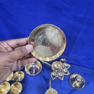 May include: A collection of brass items including a small circular plate with concentric etched rings, several small bowls, a multi-wick lamp holder with a floral shape, and a long-handled oil lamp with the text "Siddharth 2" engraved on the handle.