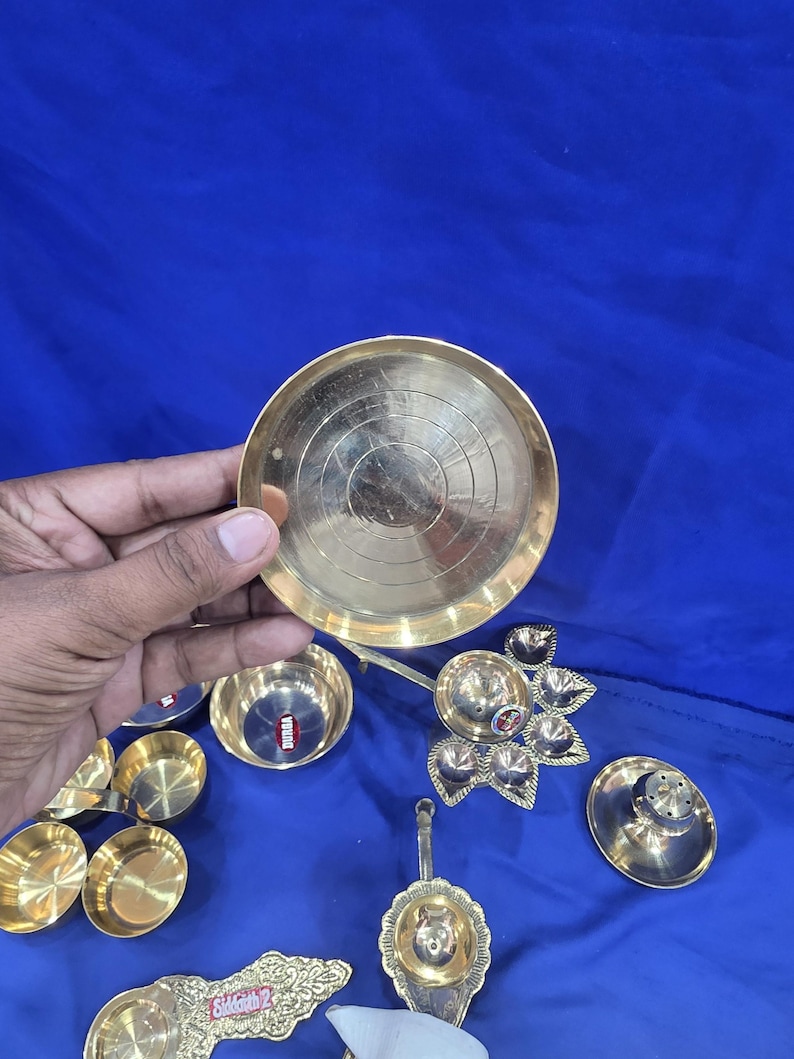 May include: A collection of brass items including a small circular plate with concentric etched rings, several small bowls, a multi-wick lamp holder with a floral shape, and a long-handled oil lamp with the text "Siddharth 2" engraved on the handle.