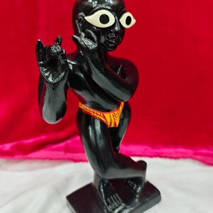 Pu&ograve; includere: Una statuetta nera di una divinit&agrave; con grandi occhi bianchi e un perizoma a fantasia gialla e rossa. La statuetta &egrave; in posa in una posizione simile alla danza, con una mano alzata e l'altra vicino al viso. Si trova su una base rettangolare nera.