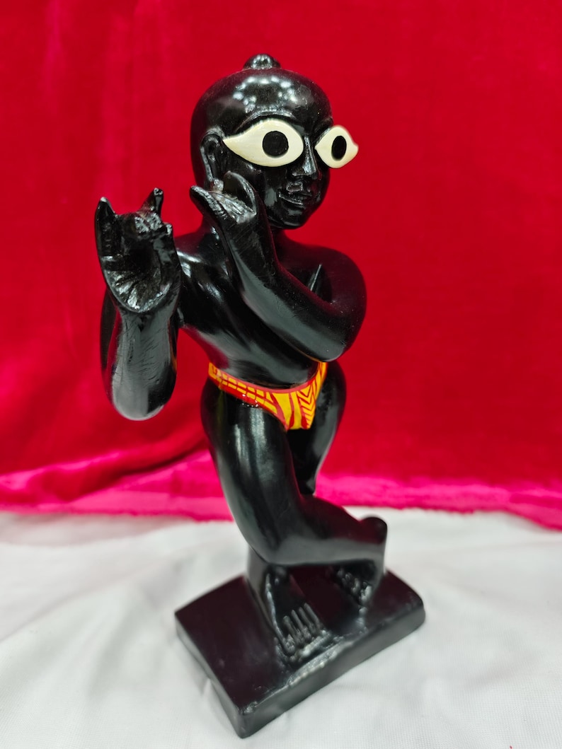 Pu&ograve; includere: Una statuetta nera di una divinit&agrave; con grandi occhi bianchi e un perizoma a fantasia gialla e rossa. La statuetta &egrave; in posa in una posizione simile alla danza, con una mano alzata e l'altra vicino al viso. Si trova su una base rettangolare nera.