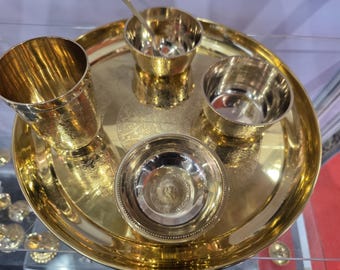 May include: A golden-colored metal thali set, including a large round tray, a tall cup, three small bowls, and a small spoon. The tray and bowls have decorative etched patterns. The set is designed for serving food.