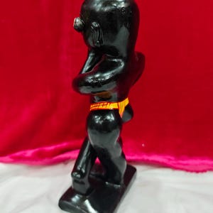 Pu&ograve; includere: Statuetta nera lucida di una persona in posizione eretta, con le braccia incrociate. La figura ha una testa piccola e indossa una fascia gialla e arancione. La statuetta &egrave; su una base rettangolare.