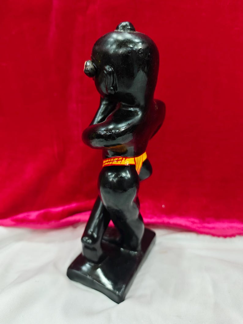 Pu&ograve; includere: Statuetta nera lucida di una persona in posizione eretta, con le braccia incrociate. La figura ha una testa piccola e indossa una fascia gialla e arancione. La statuetta &egrave; su una base rettangolare.