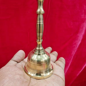 May include: A polished brass hand bell featuring a slender, vertically ribbed handle and a flared, circular base with horizontal engraved lines. The metal surface has a bright, reflective gold-toned finish, showcasing a smooth, metallic texture.