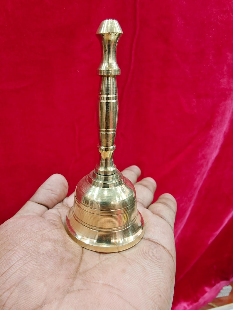 May include: A polished brass hand bell featuring a slender, vertically ribbed handle and a flared, circular base with horizontal engraved lines. The metal surface has a bright, reflective gold-toned finish, showcasing a smooth, metallic texture.