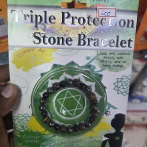 May include: A Triple Protection Stone Bracelet packaged on a green and white card. The bracelet features dark brown and tan stones. The card has text that reads "Triple Protection Stone Bracelet", "Element Air", and "Colour Green".