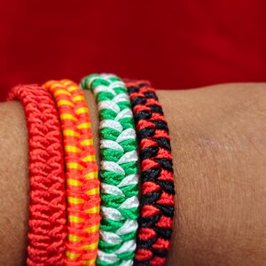 Puede incluir: Cuatro pulseras trenzadas en colores vibrantes: rojo, naranja con rayas amarillas, verde con blanco y negro con rojo. Las pulseras se llevan en una muñeca sobre un fondo rojo. Cada pulsera tiene un patrón único.