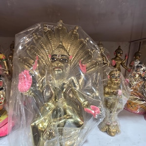 May include: A collection of gold-colored statues, including a central figure with a multi-headed serpent headdress and pink accents. Other figures are visible in the background, all appearing to be religious or mythological in theme.