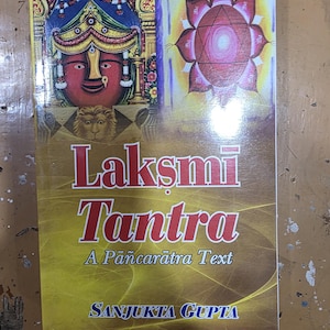 May include: A book titled "Laksmi Tantra" with a yellow and brown cover. The cover features illustrations of a deity and a lotus flower. The author's name, Sanjukta Gupta, is printed at the bottom.