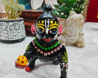 May include: A black figurine of a deity with a painted face, wearing a green and red necklace, and holding a yellow ball with red dots. The figurine has red feet and ears, and is decorated with white dots and floral designs.