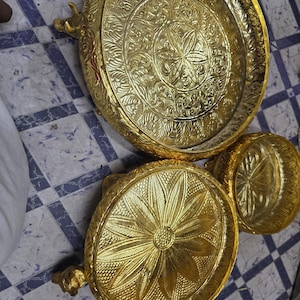 Puede incluir: Tres bandejas doradas ornamentadas con intrincados patrones florales y de hojas. Las bandejas varían en tamaño, con asas en las más grandes. Los diseños están en relieve, creando una superficie texturizada. Las bandejas están dispuestas sobre una superficie a cuadros.