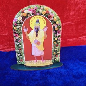 May include: A decorative arch-shaped stand featuring an image of a religious figure in yellow robes, with a long beard and raised hands. The image is framed by a floral arch of colorful roses and other flowers. The base is green, and the background is red and blue.