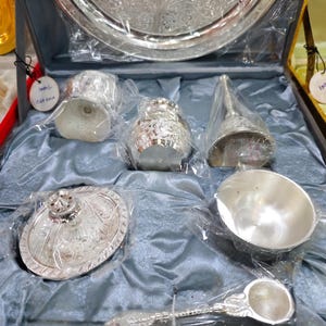 May include: A silver-colored serving set displayed in a dark blue box. The set includes a large tray, small cups, a bell, a bowl, a spoon, and a covered dish. Each piece is intricately designed with floral patterns and is wrapped in clear plastic.