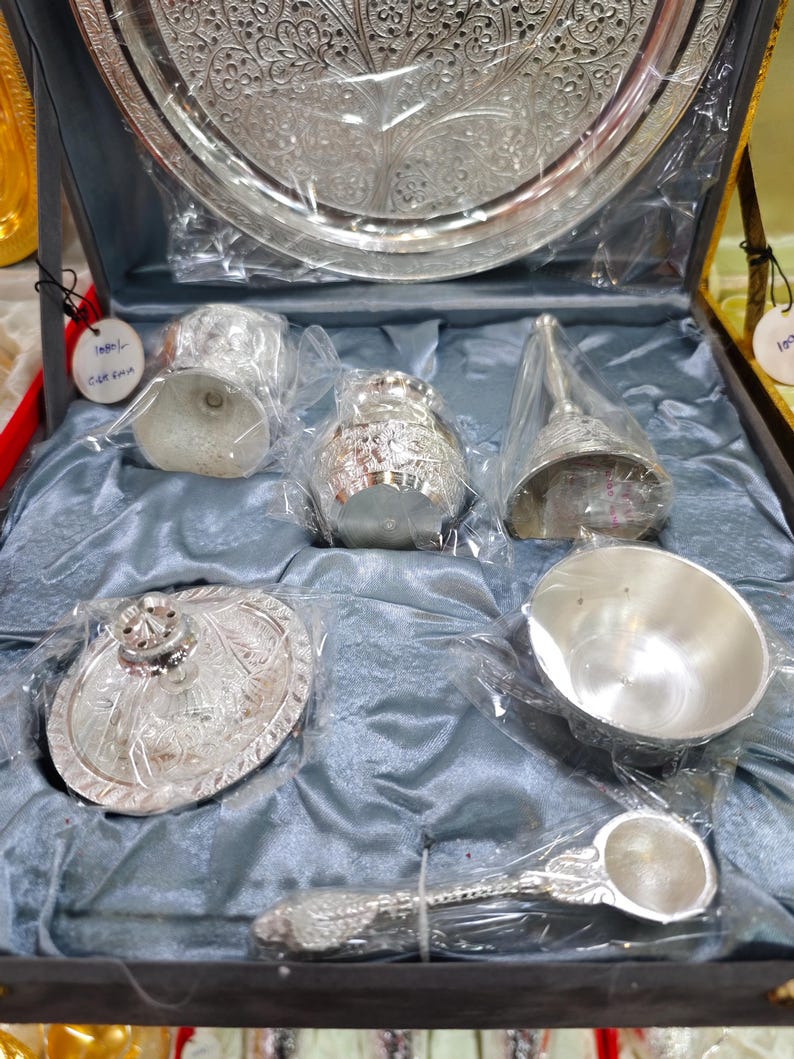 May include: A silver-colored serving set displayed in a dark blue box. The set includes a large tray, small cups, a bell, a bowl, a spoon, and a covered dish. Each piece is intricately designed with floral patterns and is wrapped in clear plastic.