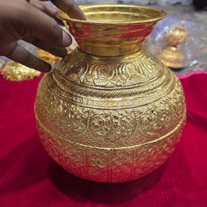 May include: A gold-colored, ornate metal pot with a wide, flared rim and intricate embossed patterns. The pot has a rounded body and a smaller, cylindrical neck. The design features detailed floral and geometric motifs.