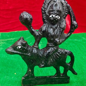May include: A black cast iron sculpture of a deity riding a bull. The deity holds a ball in one hand and a curved object in the other. The bull has white eyes. The sculpture is on a green base with a red and green background.
