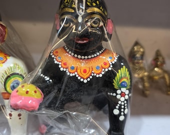 May include: A black figurine of a deity, adorned with gold and colorful accents. The figure is wrapped in clear plastic and features intricate details on the face and clothing. The figurine is part of a collection of religious figures.