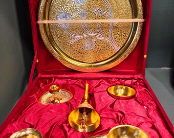 May include: A gold-colored decorative set with a large round tray featuring a floral design in silver and gold. The set includes a bell, small bowls, and a decorative pot, all displayed in a red velvet-lined box.