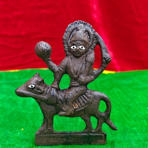 May include: A dark brown cast iron statue of a deity riding an animal, possibly a bull. The deity holds a sphere in one hand. The statue has white painted eyes and is set against a red and green background.