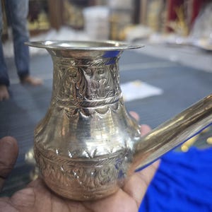 May include: A silver-colored metal water vessel with a spout and ornate, engraved floral and figure designs. The vessel has a wide, flared rim and a bulbous body. The spout extends from the side.