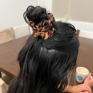 May include: A scrunchie with a brown and black leopard print is worn in the hair. The scrunchie is made of a soft fabric and is tied around a bun. The hair is dark and long. The background includes a table and a person holding a mug.