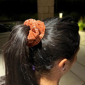 May include: A scrunchie with a plaid pattern in shades of orange and white is worn in a ponytail. The hair is dark and straight. The scrunchie adds a pop of color to the hairstyle. The background is blurred, with hints of a building and greenery.