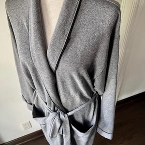May include: A gray robe with a shawl collar, long sleeves, and a tie belt. The robe has two front pockets and black trim along the edges. The fabric appears to be a soft knit material.