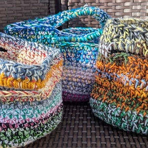 May include: Three handmade crochet baskets in various sizes and colours. The baskets feature a mix of blue, green, yellow, orange, and purple yarn. Each basket has a sturdy handle, perfect for carrying small items or as decorative storage.
