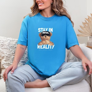 Könnte beinhalten: Ein hellblaues T-Shirt mit dem Text "STAY IN VIRTUAL REALITY" und einer Grafik eines Teddybären mit VR-Brille. Der Satz "Where dreams happen" ist unter dem Wort "REALITY" gedruckt.