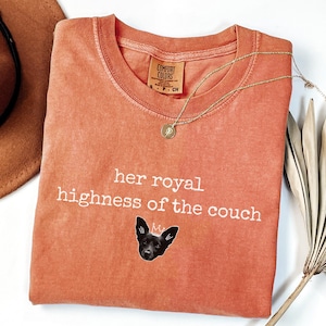 May include: An orange Comfort Colors t-shirt with the text "her royal highness of the couch" and a graphic of a dog wearing a crown. A gold necklace with a pendant is draped across the shirt.