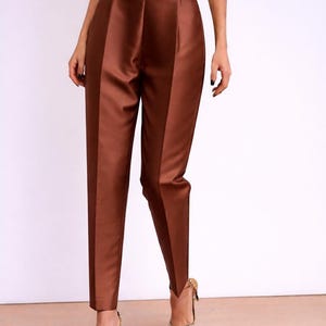 May include: Brown satin trousers with a high waist and tailored fit. The trousers have a subtle sheen and are paired with gold-coloured heels adorned with embellishments. The overall look is elegant and sophisticated.