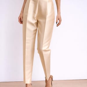 May include: Cream-coloured high-waisted trousers with a tailored fit. The trousers have a slight sheen and a wide waistband. The model is wearing patterned heels. The trousers are a neutral colour, suitable for various occasions.