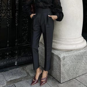 May include: Black high-waisted trousers with a vertical black stripe down the side. The trousers are paired with burgundy slingback heels with buckle details. The outfit is completed with a black top.