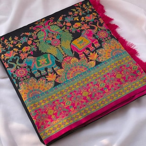 K&ouml;nnte beinhalten: Gefalteter schwarzer Stoff mit lebendigen Blumen- und Tiermotiven. Das Design zeigt Elefanten, Pfauen und komplizierte Muster in Rosa-, Gelb-, T&uuml;rkis- und Oranget&ouml;nen. Der Rand ist mit einem rosa Fransenbesatz versehen.