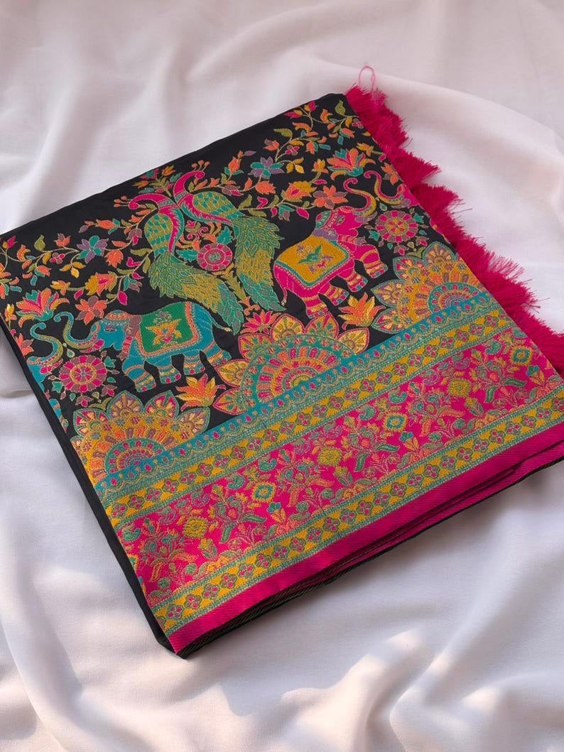 K&ouml;nnte beinhalten: Gefalteter schwarzer Stoff mit lebendigen Blumen- und Tiermotiven. Das Design zeigt Elefanten, Pfauen und komplizierte Muster in Rosa-, Gelb-, T&uuml;rkis- und Oranget&ouml;nen. Der Rand ist mit einem rosa Fransenbesatz versehen.