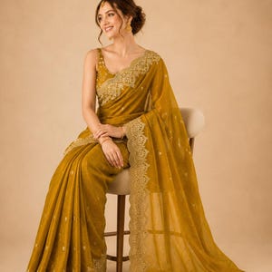 May include: Mustard yellow saree with intricate gold embroidery along the borders and neckline. The saree is draped elegantly, showcasing its flowing fabric and detailed embellishments. The outfit includes a matching sleeveless blouse.