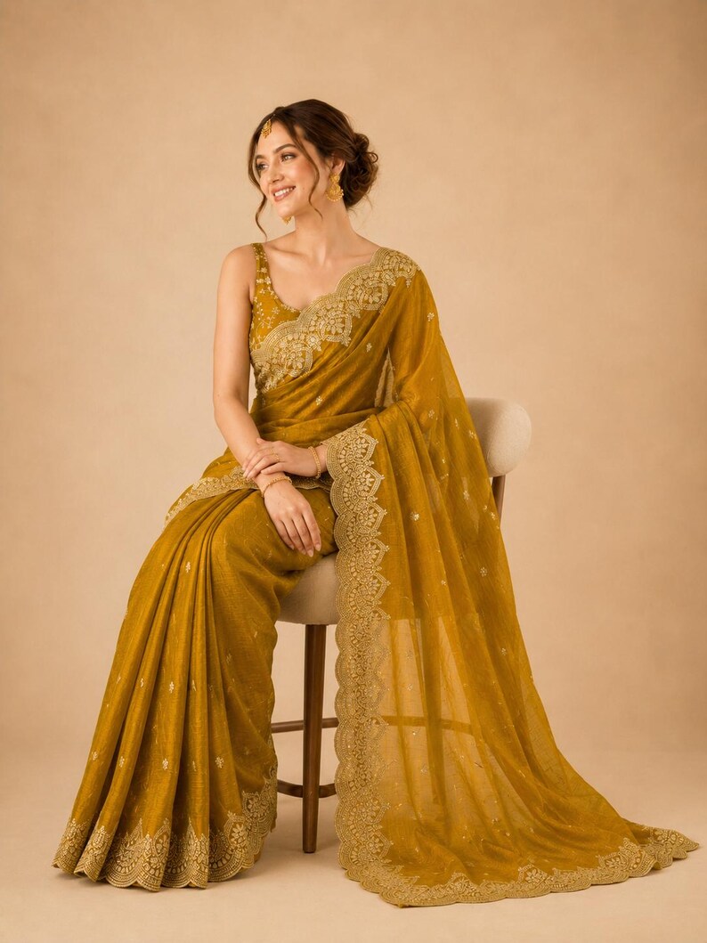 May include: Mustard yellow saree with intricate gold embroidery along the borders and neckline. The saree is draped elegantly, showcasing its flowing fabric and detailed embellishments. The outfit includes a matching sleeveless blouse.