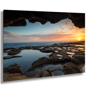 Puede incluir: Fotografía de paisaje de una escena costera al atardecer. La imagen captura una vista desde el interior de una formación rocosa, mirando hacia el océano. El cielo está lleno de tonos naranjas y rosas, y el agua refleja los colores del atardecer.