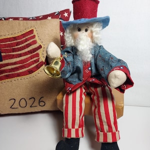 May include: A patriotic Uncle Sam doll with a red, white, and blue color scheme. The doll wears a blue coat with a star pattern, red and white striped pants, and a red top hat. It holds a small gold bell. A pillow with an American flag design is in the background.