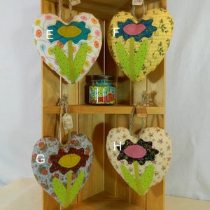 May include: Five handmade heart-shaped pillows with embroidered flowers. Each pillow has a different fabric pattern and flower design. The pillows are displayed on a wooden shelf.