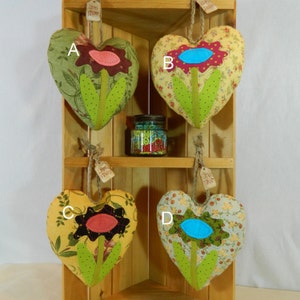 May include: Four heart-shaped fabric ornaments with floral designs. Each heart features a different color flower with a green stem and leaves. The hearts are hanging from a wooden shelf.