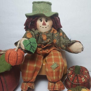 May include: A fabric scarecrow doll with a green hat, red yarn hair, and a stitched face. It wears a green and orange pumpkin-patterned shirt and orange plaid pants. The doll is holding a small pumpkin and a green leaf. Other decorative pumpkins are nearby.