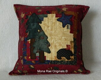 May include: A red, green, and blue quilted pillow with a bear, pine trees, and a star design. The pillow has the text "Mona Rae Originals ©" on the bottom.