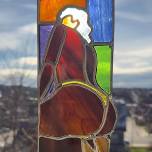 May include: Stained glass panel depicting a figure in a seated position, with a white head and red robe. The panel features a rectangular shape with colorful glass sections in orange, blue, green, purple, and yellow. The artwork is suspended by a metal chain.