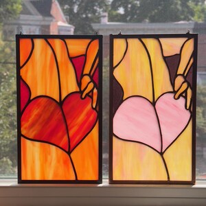 May include: Two stained glass panels with black frames. One features a red heart shape, the other a pink heart. Both have yellow and orange backgrounds, with a hand reaching towards the heart.