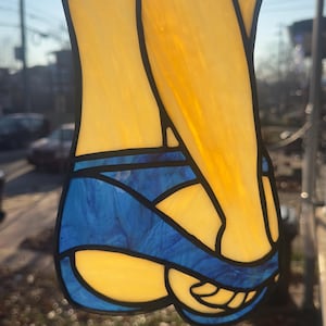May include: Stained glass suncatcher depicting a figure in a swimsuit. The design features yellow and blue glass, with black outlines. The suncatcher is suspended by a silver chain. The overall shape is abstract and artistic.