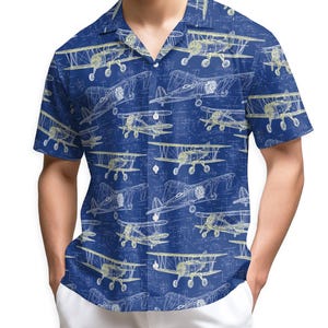 May include: A short-sleeved, button-down shirt with a navy blue background and a repeating pattern of vintage airplane blueprints in white and yellow. The shirt has a classic collar and is worn by a person.