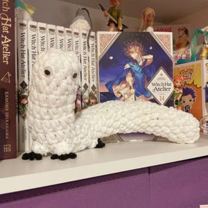 Puede incluir: Un gusano blanco de ganchillo hecho a mano, con ojos saltones y pies negros. El gusano está colocado en un estante frente a una fila de libros y figuras. Los libros se titulan "Witch Hat Atelier".