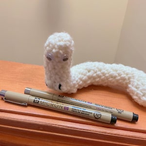 Puede incluir: Un gusano de ganchillo blanco hecho a mano con ojos saltones y dos bolígrafos Micron beige. Los bolígrafos están etiquetados como "Micron 03" y "Micron 08". El gusano está sobre una superficie de madera.