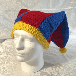 May include: A handmade crochet jester hat in red, blue, and yellow. The hat features a pointed top with a yellow pom-pom and a yellow brim. The hat is displayed on a white mannequin head.